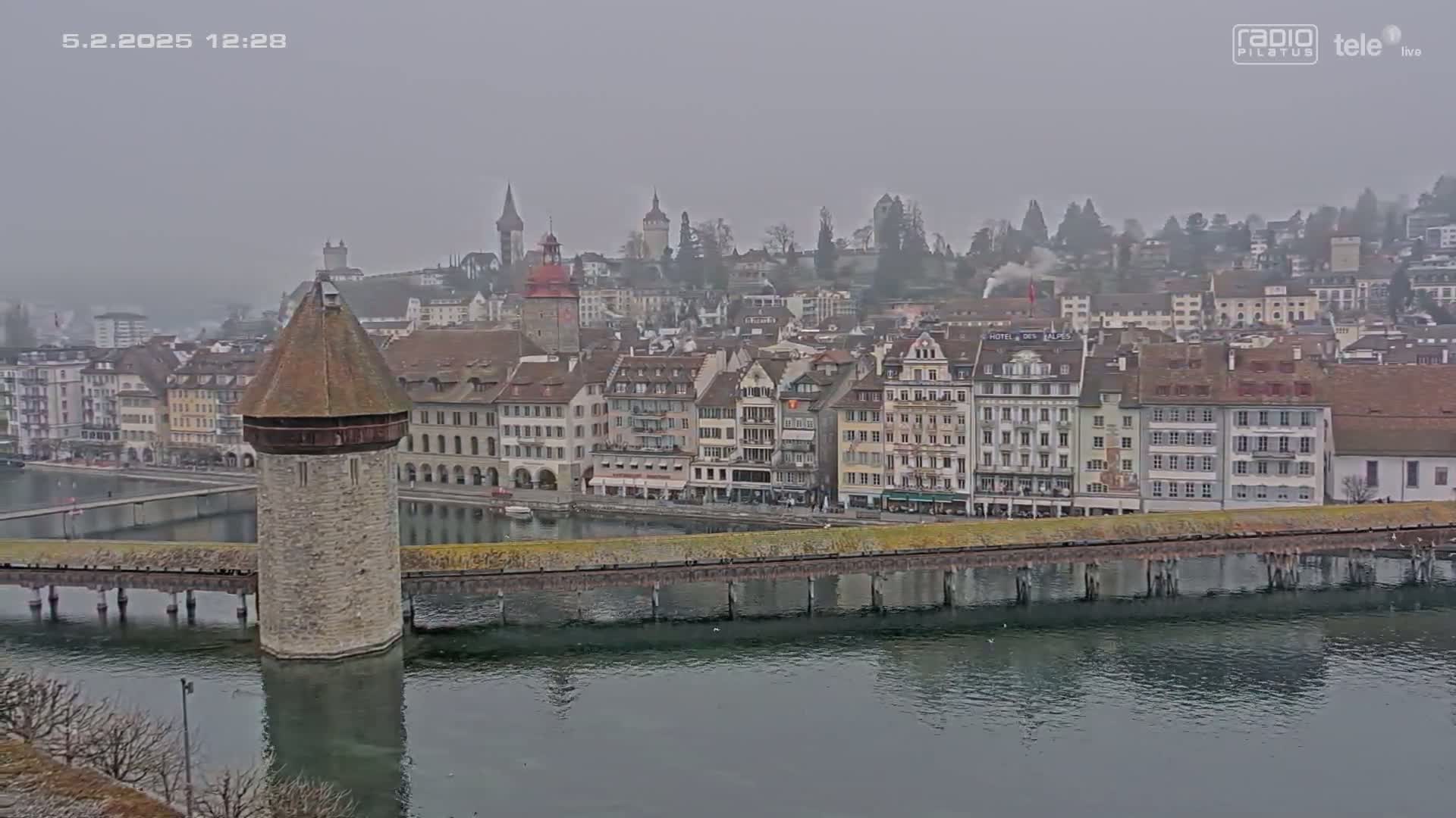 A stone water tower stands on a bridge over a calm river, with a foggy cityscape of multi-story buildings behind it.