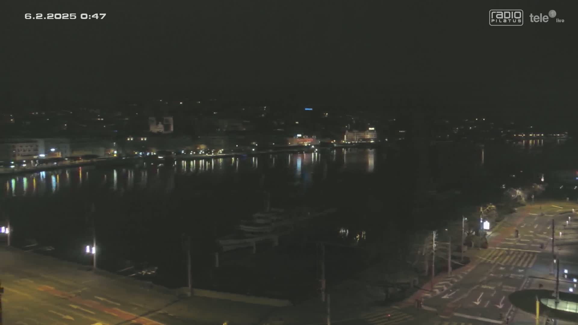 A nighttime view shows a city's waterfront with calm water reflecting the lights of buildings and streets, under a clear, dark sky.