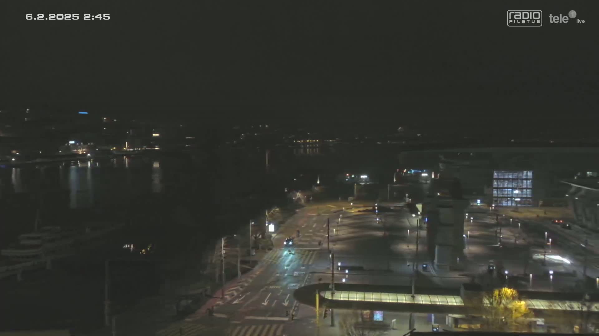 A nighttime view of a city waterfront with a calm body of water, sparsely lit streets, and a mostly empty parking lot.