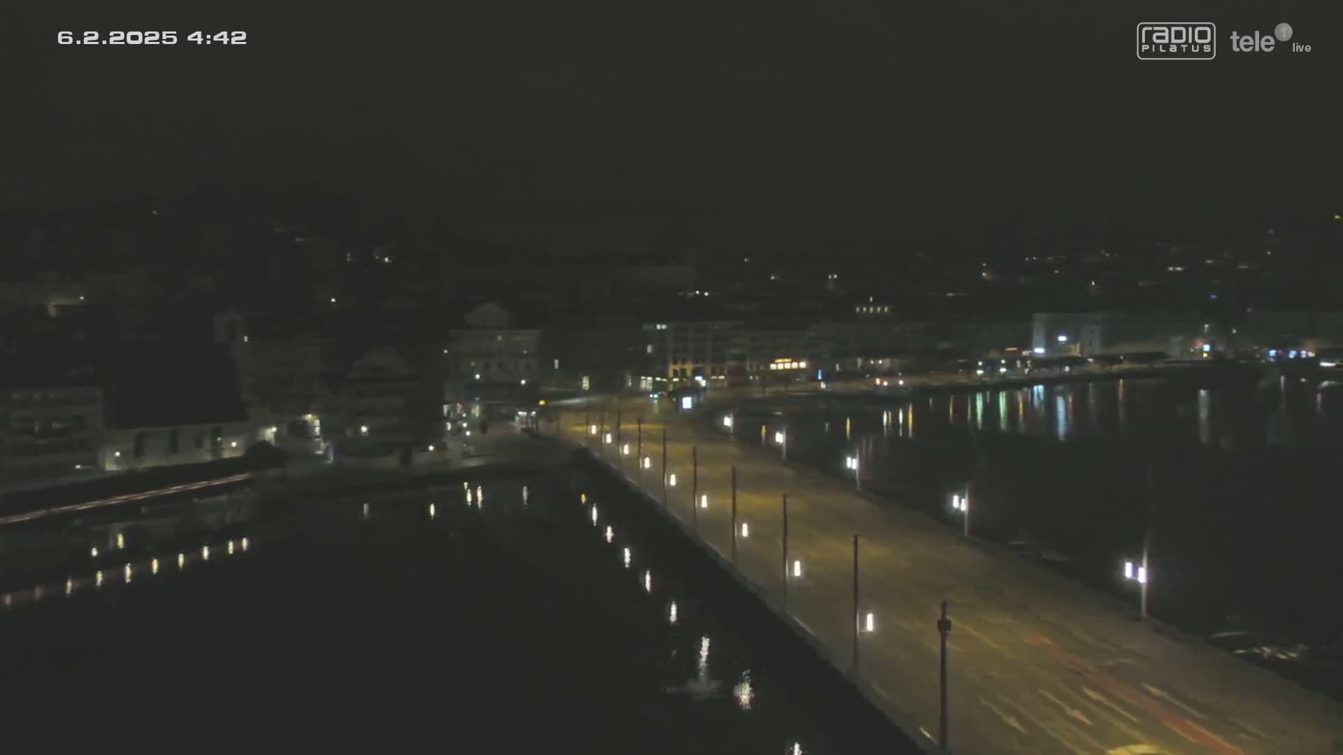 A nighttime, long shot of a city waterfront with a roadway lined with lights, reflecting in calm water.