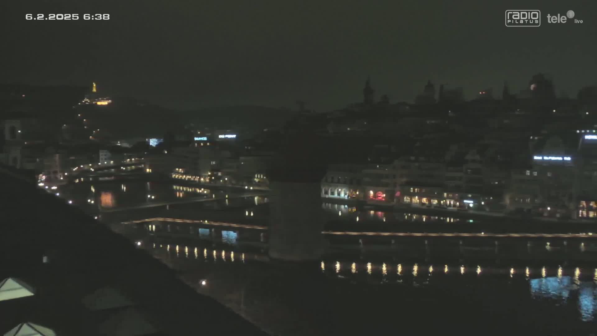 A nighttime, long shot of a city beside a calm river, with dimly lit buildings and a hill in the background.