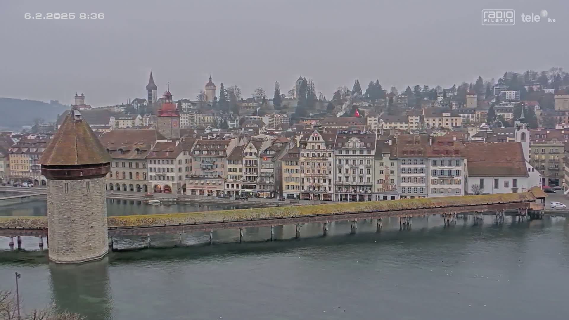 A stone tower and covered wooden bridge extend over a body of water in front of a town of European-style buildings under an overcast sky.