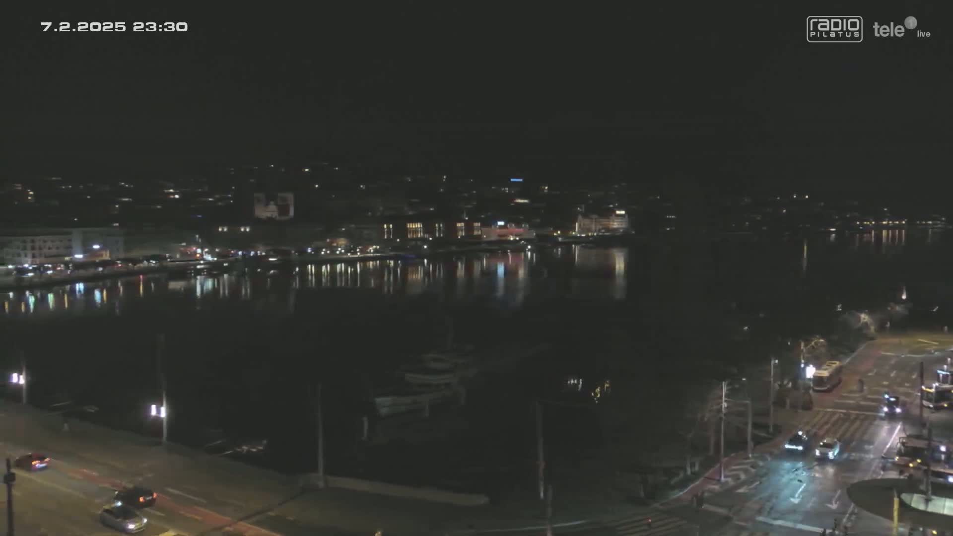 A nighttime view of a city waterfront, with lights reflecting on a calm body of water, and cars driving along a road in the foreground.