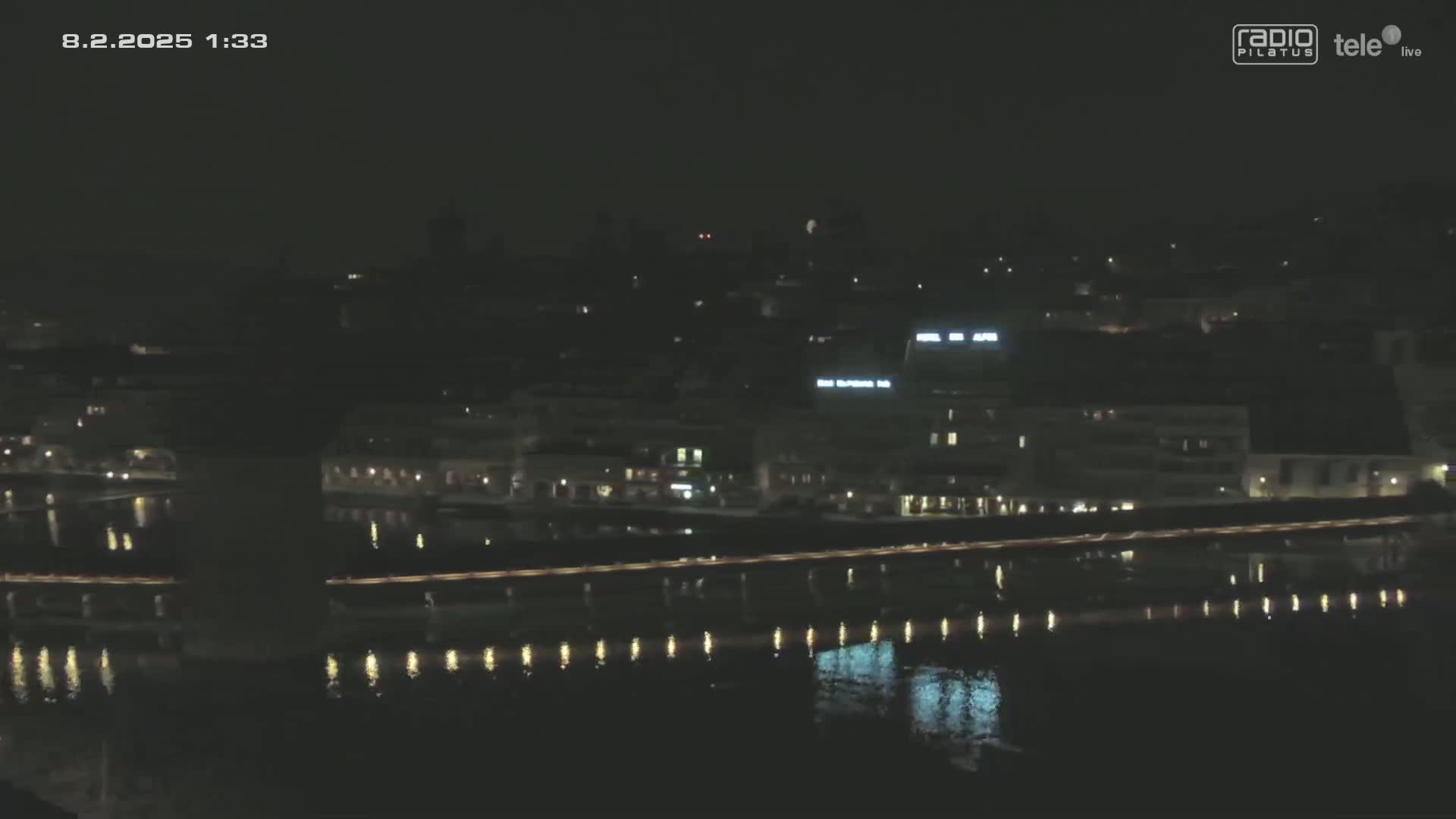 A nighttime long shot shows a city waterfront at night with calm water reflecting city lights.