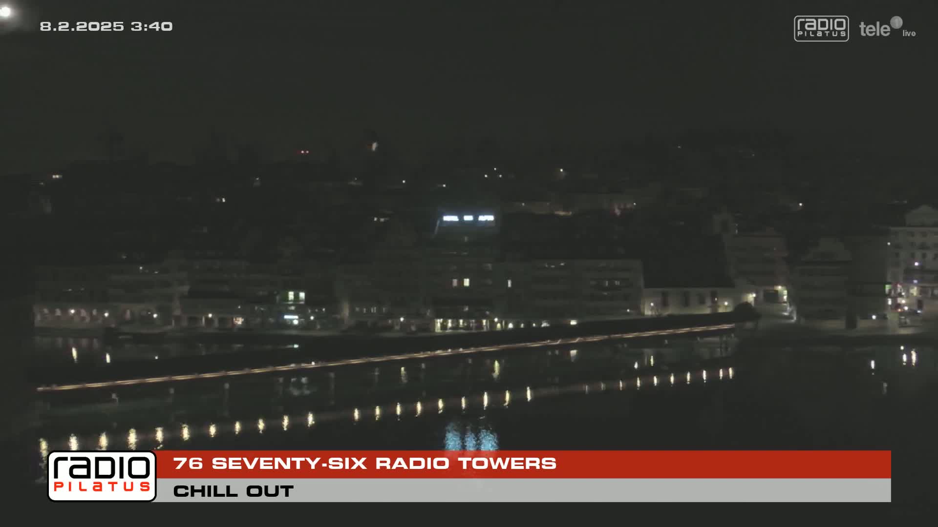 A nighttime view of a city waterfront with calm water reflecting city lights under a clear sky.