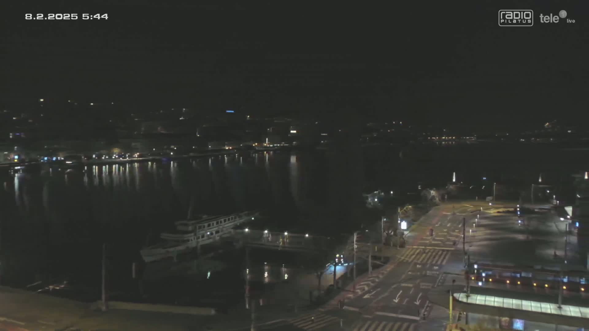 A nighttime, high-angle view shows a body of water reflecting city lights, a docked boat, and a roadway with some traffic.