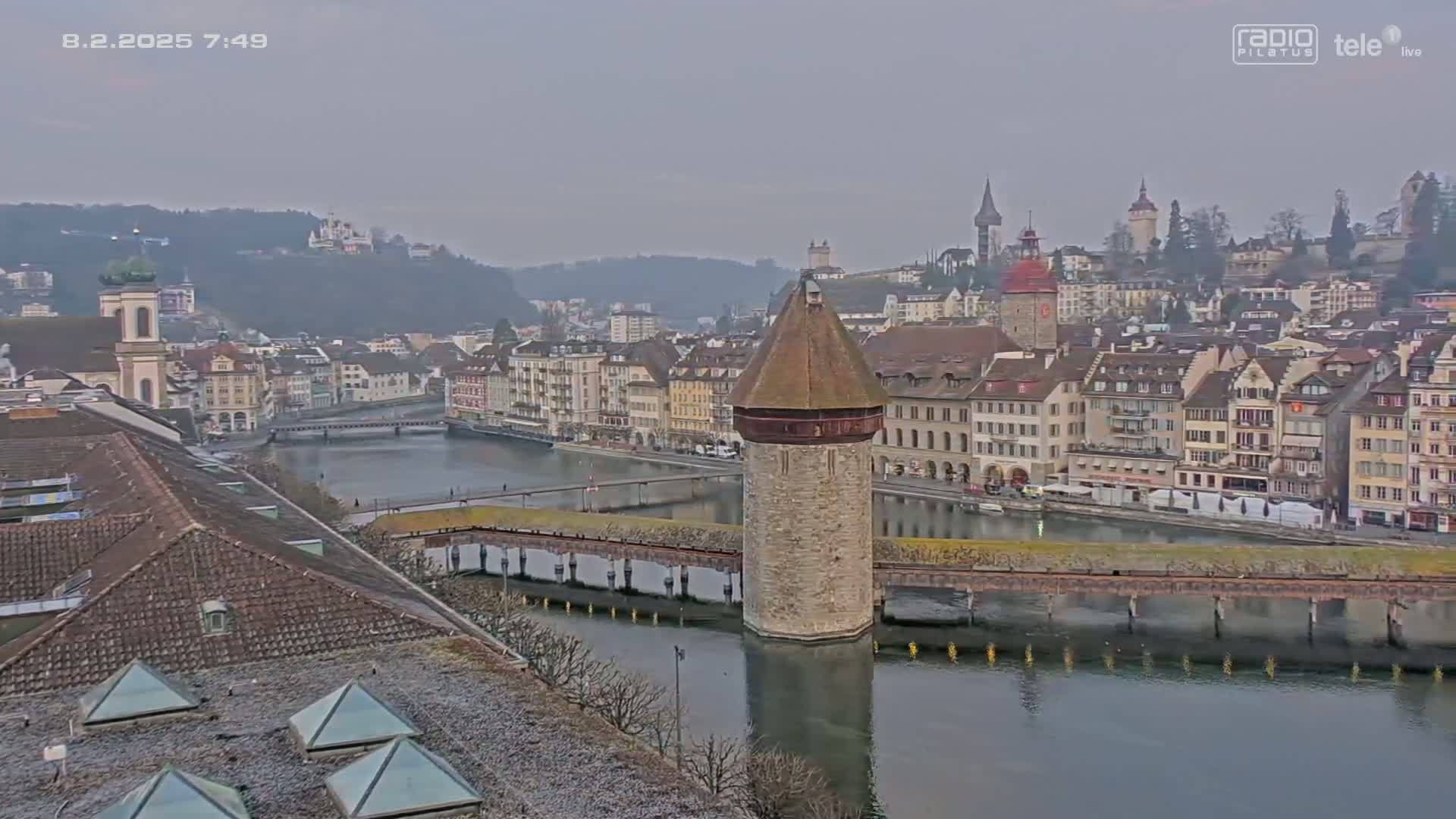 A stone water tower stands on a bridge over a calm river, surrounded by a European-style city on a cloudy day.