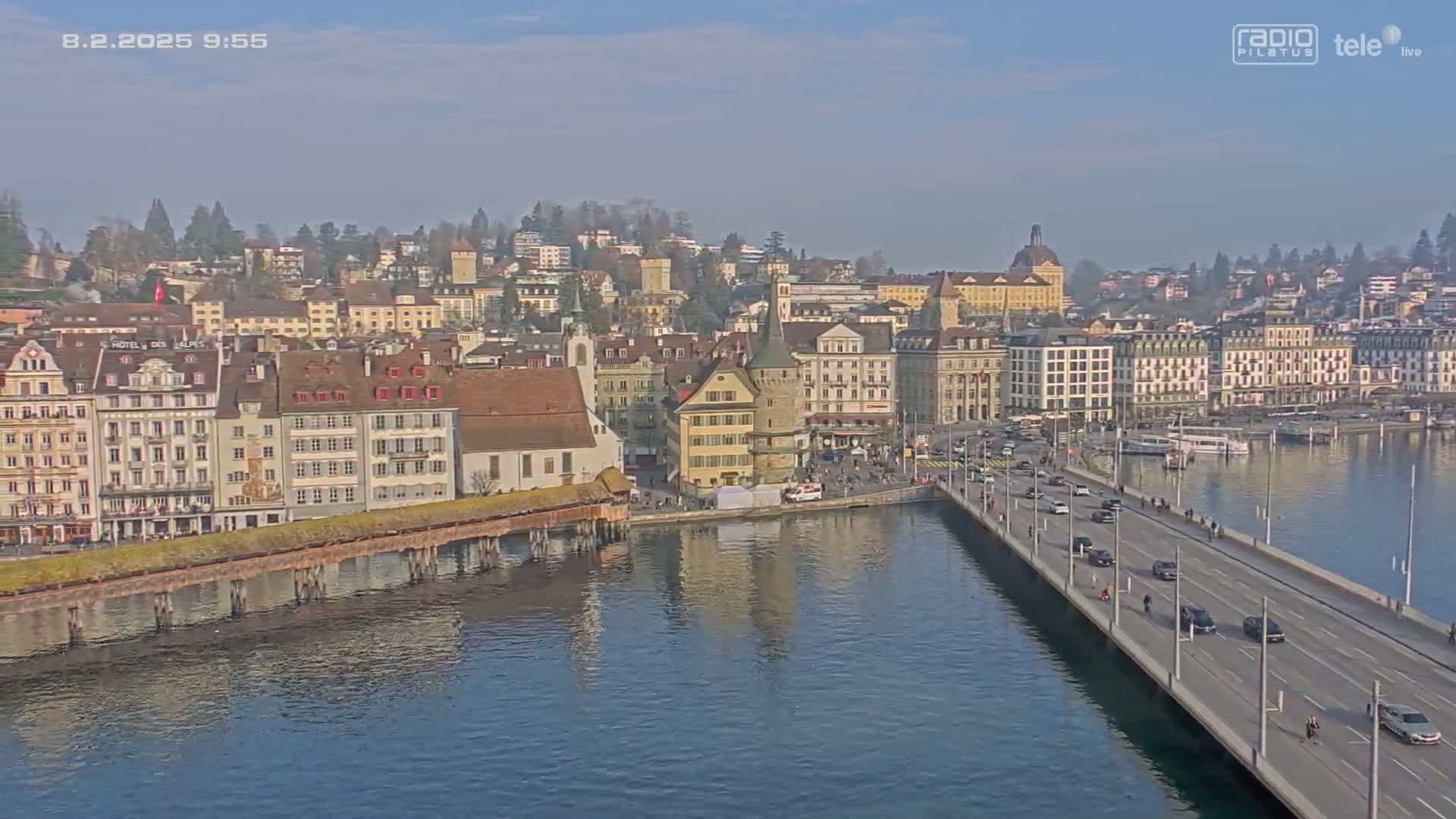 A bridge stretches across a calm body of water, leading to a city of historic buildings under a clear, sunny sky.