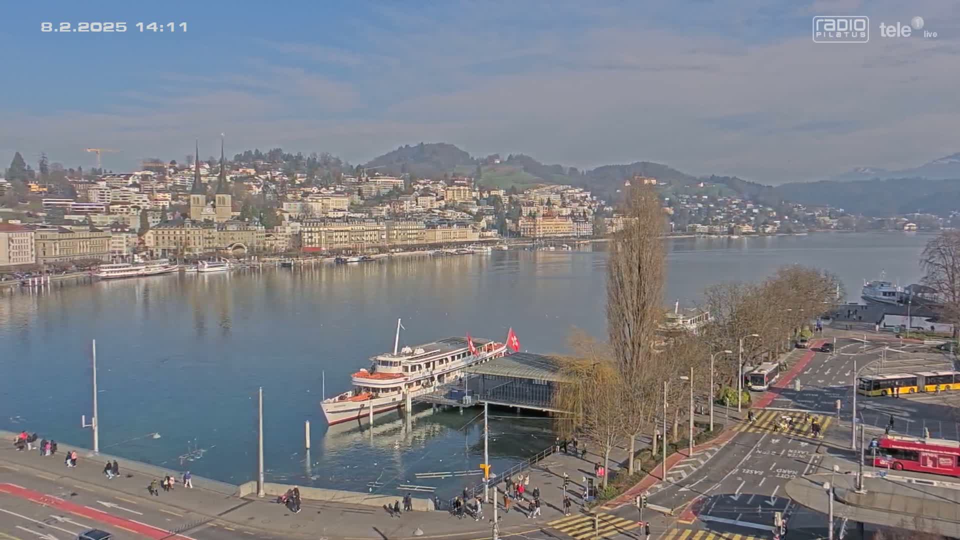 A lakeside city with a docked passenger ship is seen on a sunny day, with clear skies and calm water.