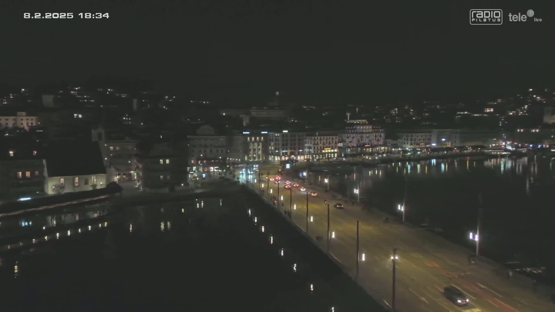 A nighttime aerial view of a city waterfront, showing a calm body of water reflecting the lights of buildings and a road with sparse traffic.