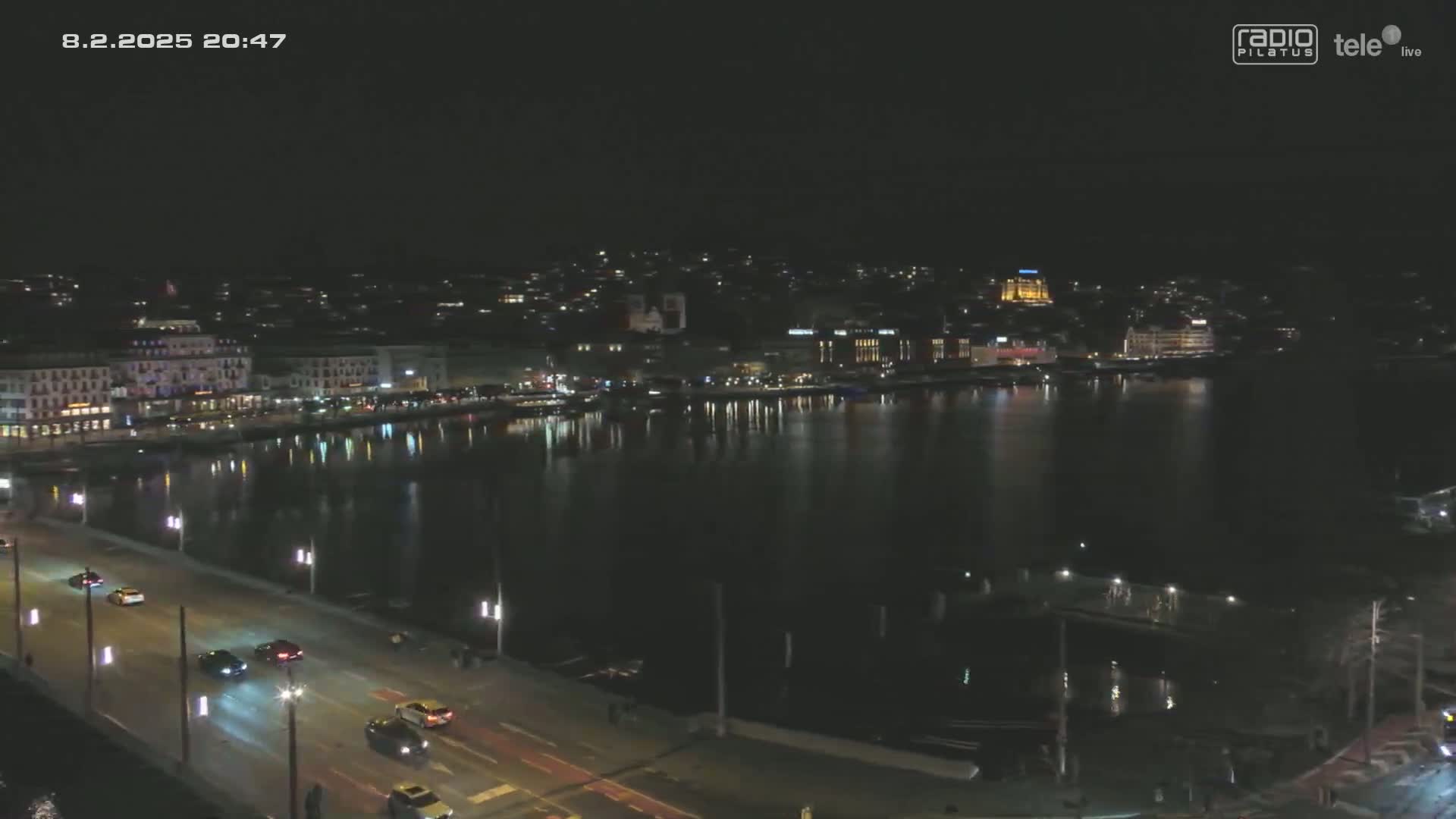 A nighttime view of a city waterfront with a calm body of water reflecting the city lights, and a road with cars driving along it.