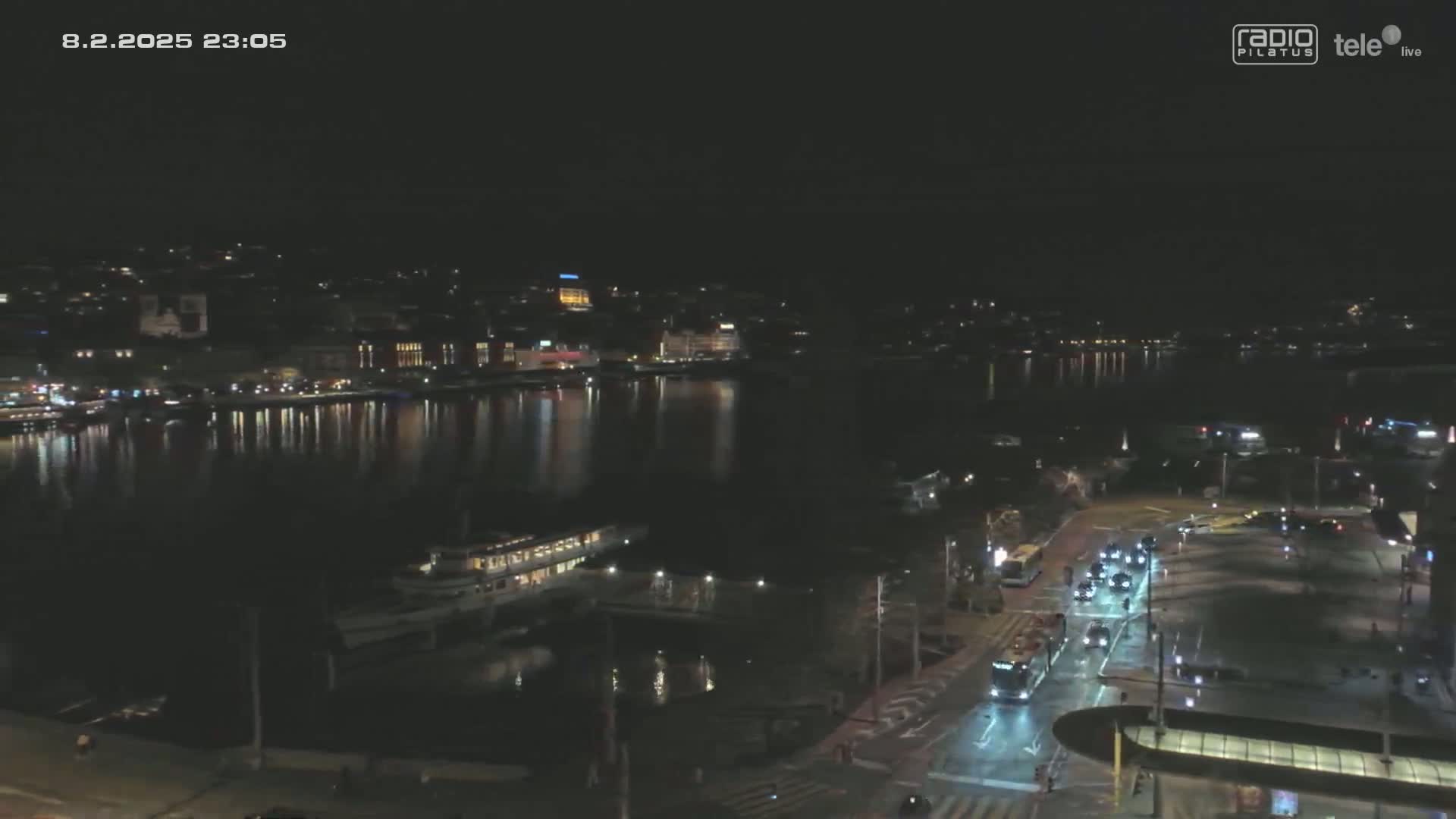 A nighttime view of a city waterfront, featuring a calm body of water reflecting city lights, boats docked at the shore, and a roadway with vehicles.