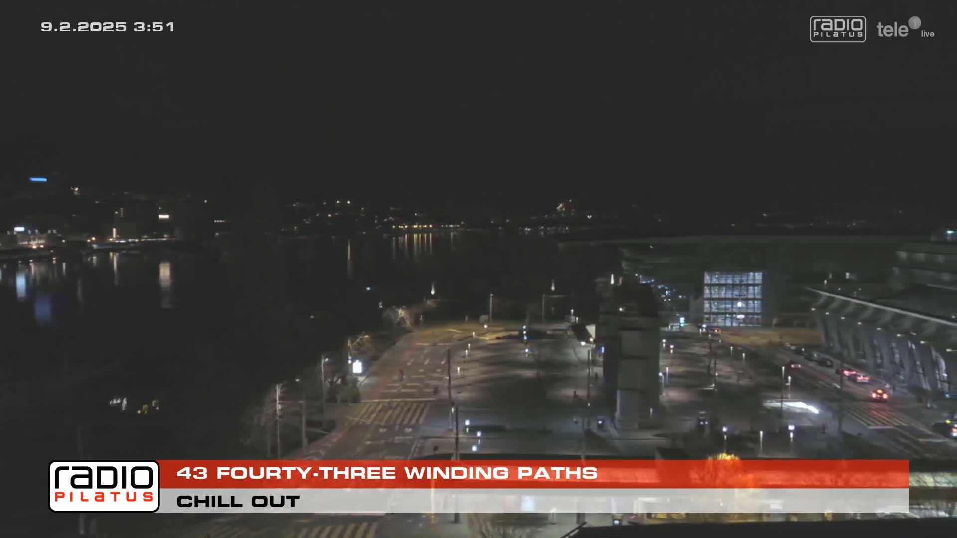 A nighttime, wide shot of a city waterfront with sparsely trafficked roads and a body of dark water reflecting city lights.