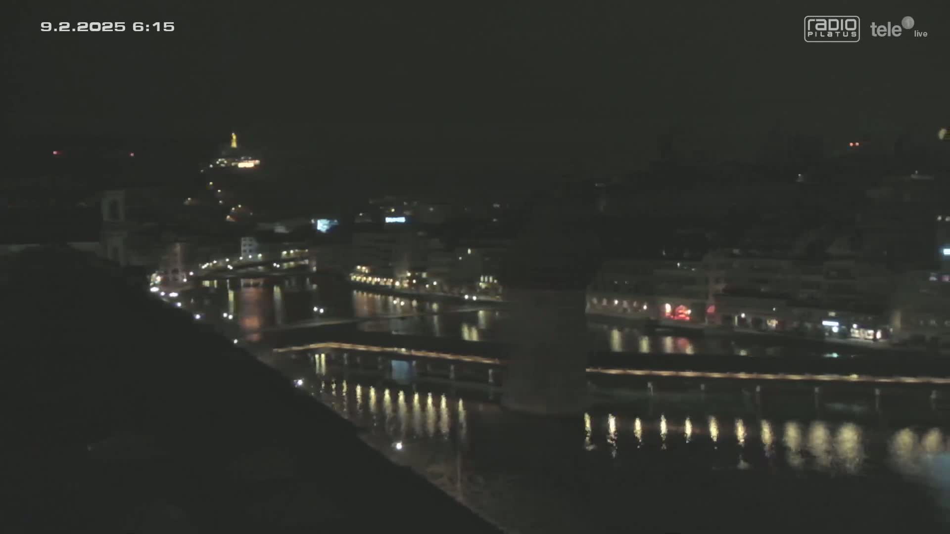 A nighttime view of a city with a river reflecting lights from buildings along its banks under a clear, dark sky.