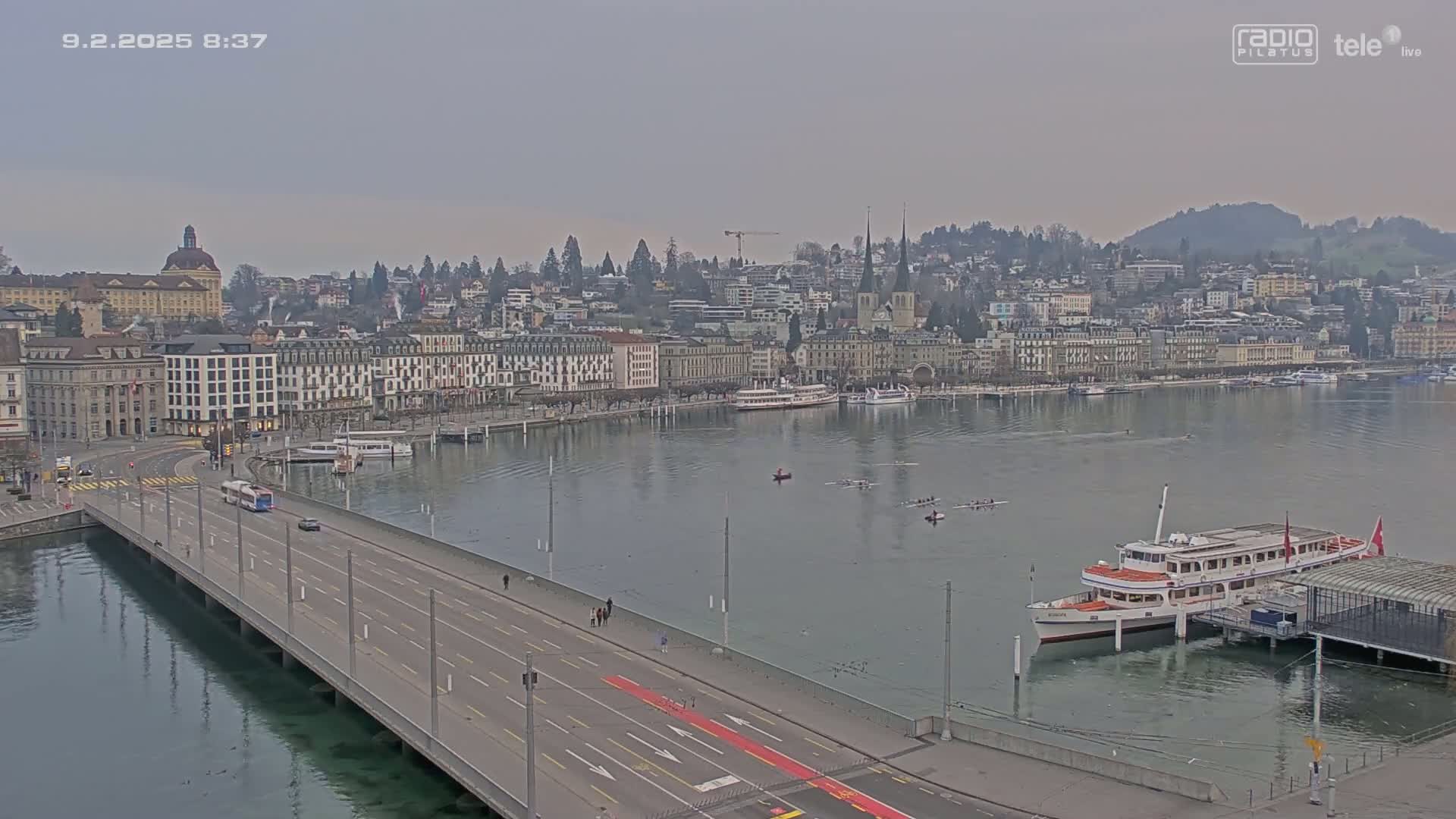 A long bridge over calm water leads to a city of buildings along a waterfront, with several boats and people rowing in the water under an overcast sky.