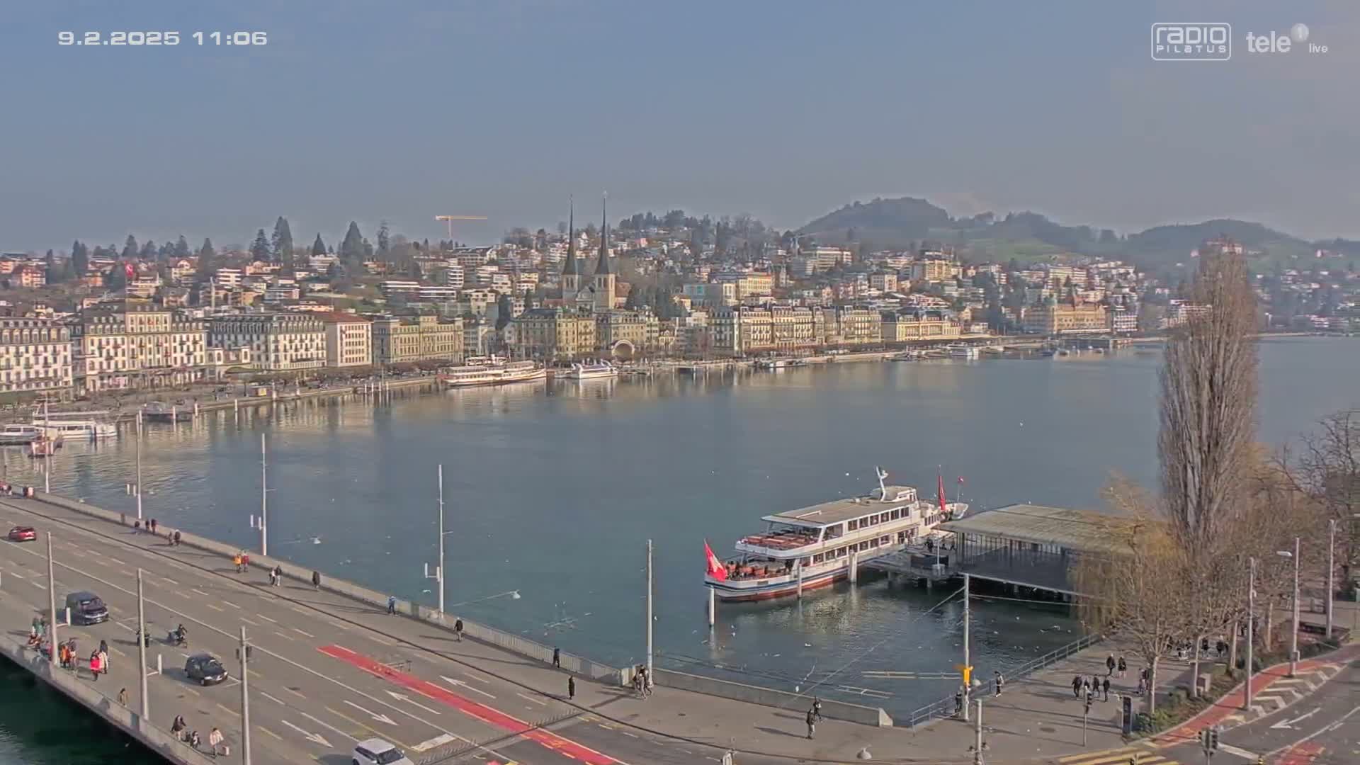 A city waterfront on a sunny day shows a bridge with cars and pedestrians, a docked passenger boat, and buildings along the water's edge.