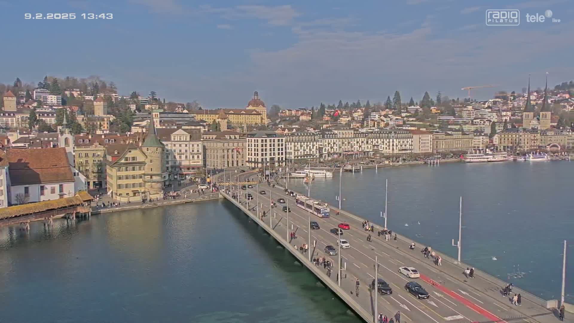 A bridge spans a body of water in front of a city on a sunny day, with boats docked at the waterfront.