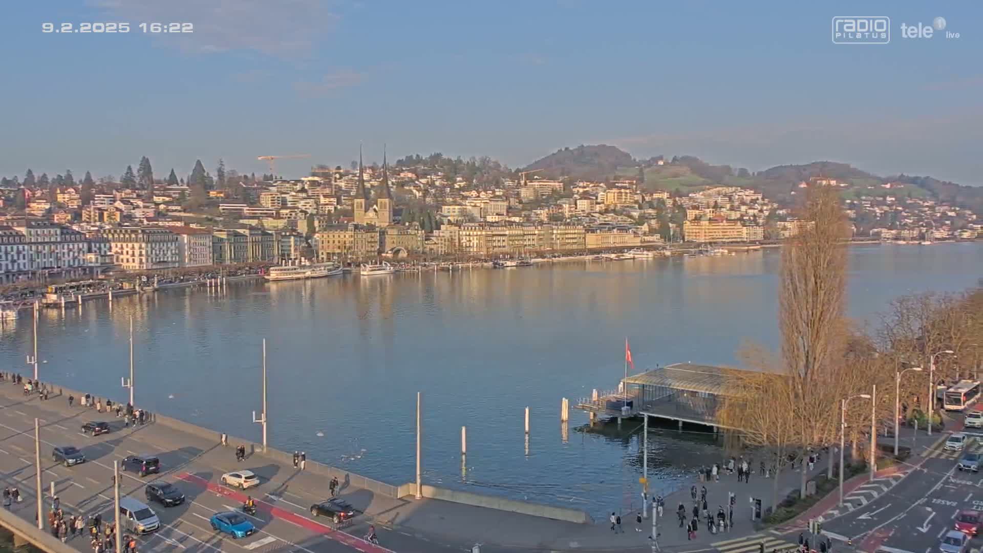 A clear, sunny day overlooks a city waterfront with buildings lining the shore of a calm lake, a bridge with cars driving on it, and people walking along the sidewalks.