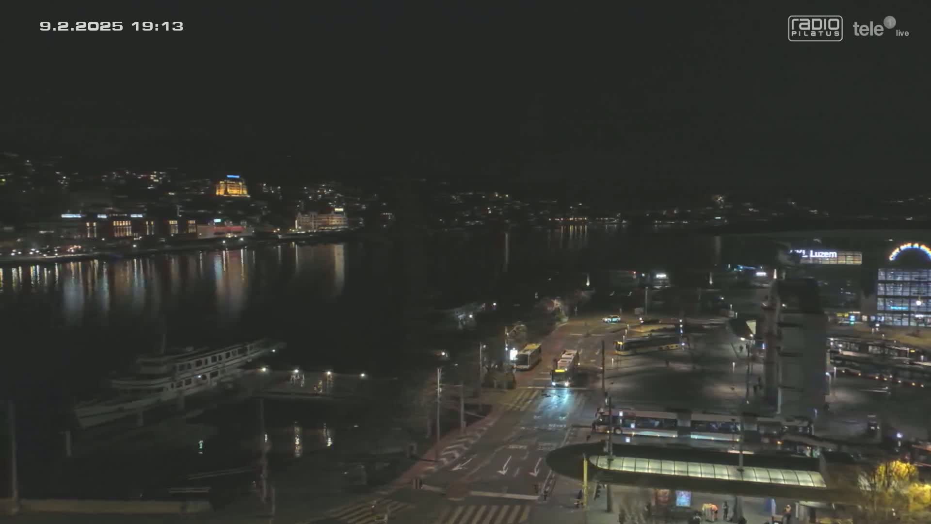 A nighttime view of a city harbor with calm water reflecting city lights, several buses and trams at a transit station, and a large building in the background.
