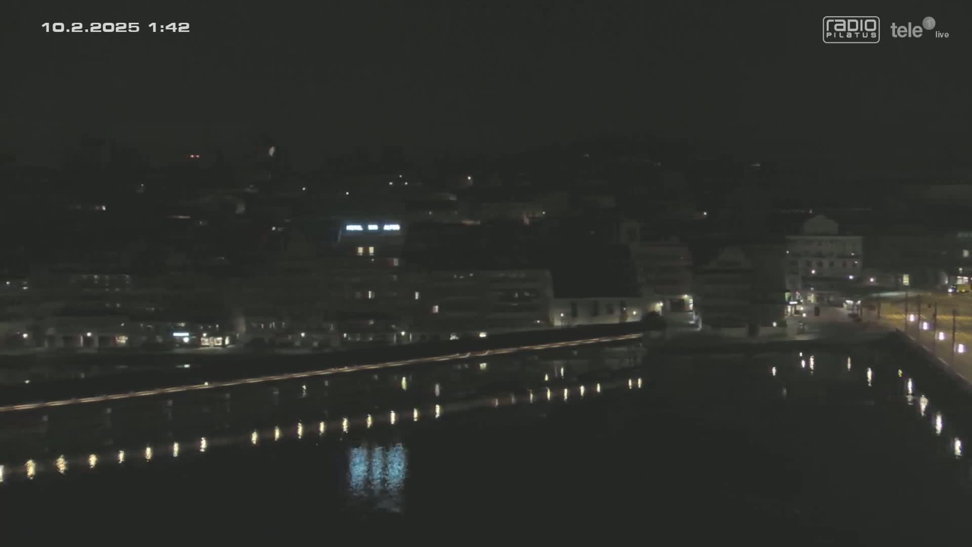 A nighttime view of a city's waterfront, showing buildings and lights reflected in dark water under a clear sky.