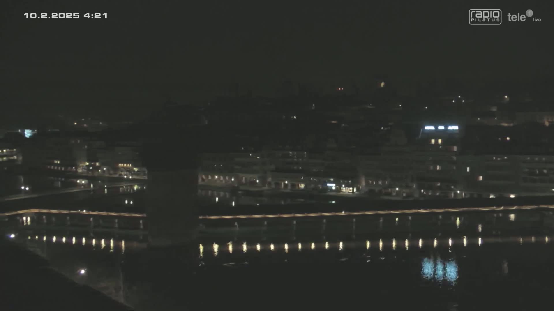 A nighttime view of a city waterfront, showing buildings with lights reflecting on a dark body of water under a clear, dark sky.