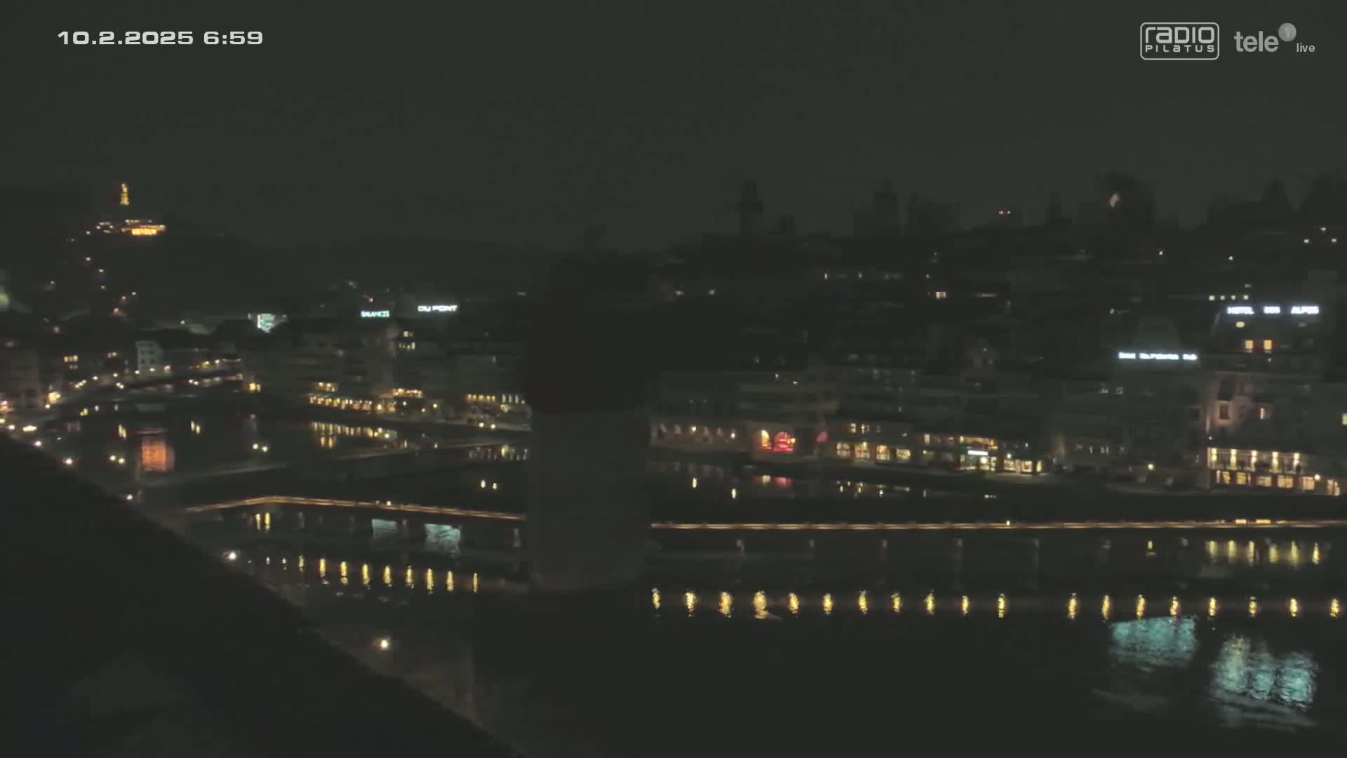 A nighttime view of a city with buildings and lights reflected in a calm body of water under a dark sky.
