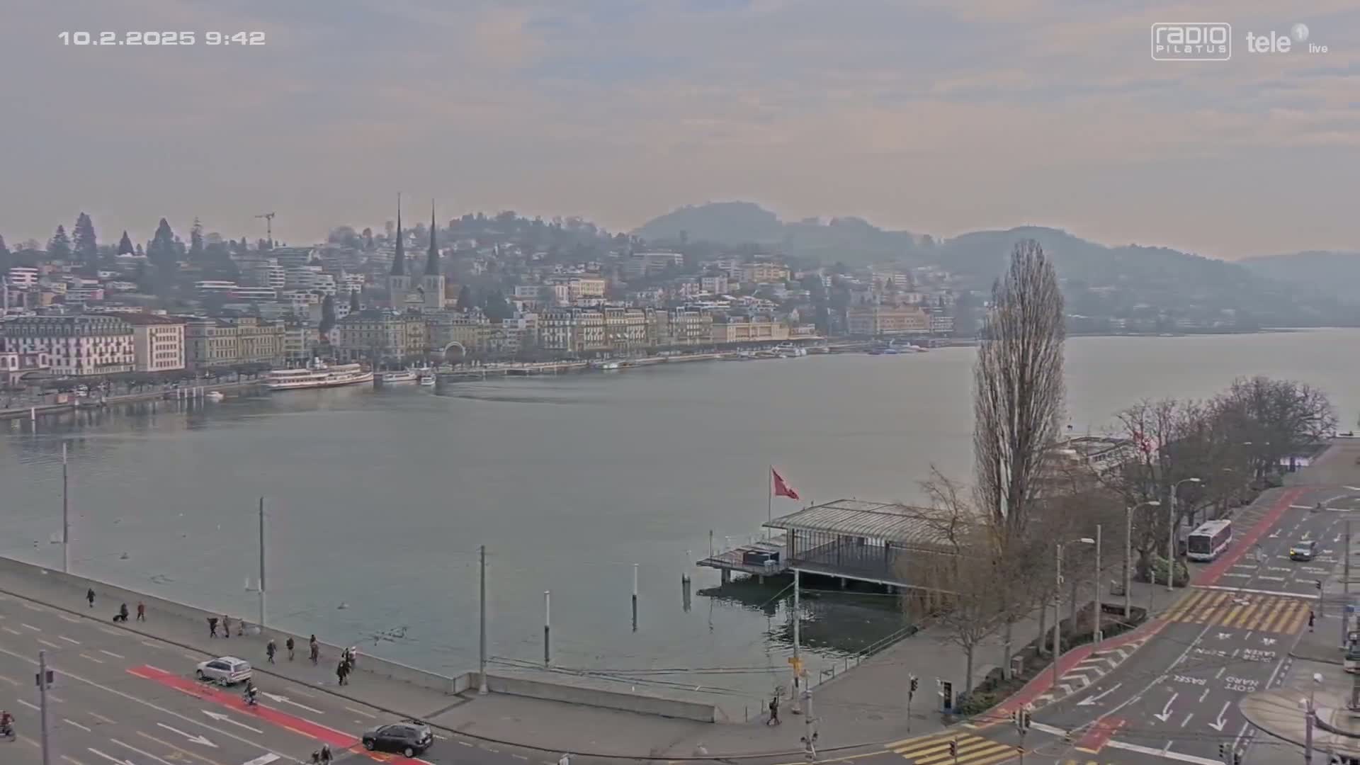 A hazy view of a city situated on a lake, with a street and some pedestrians visible in the foreground.