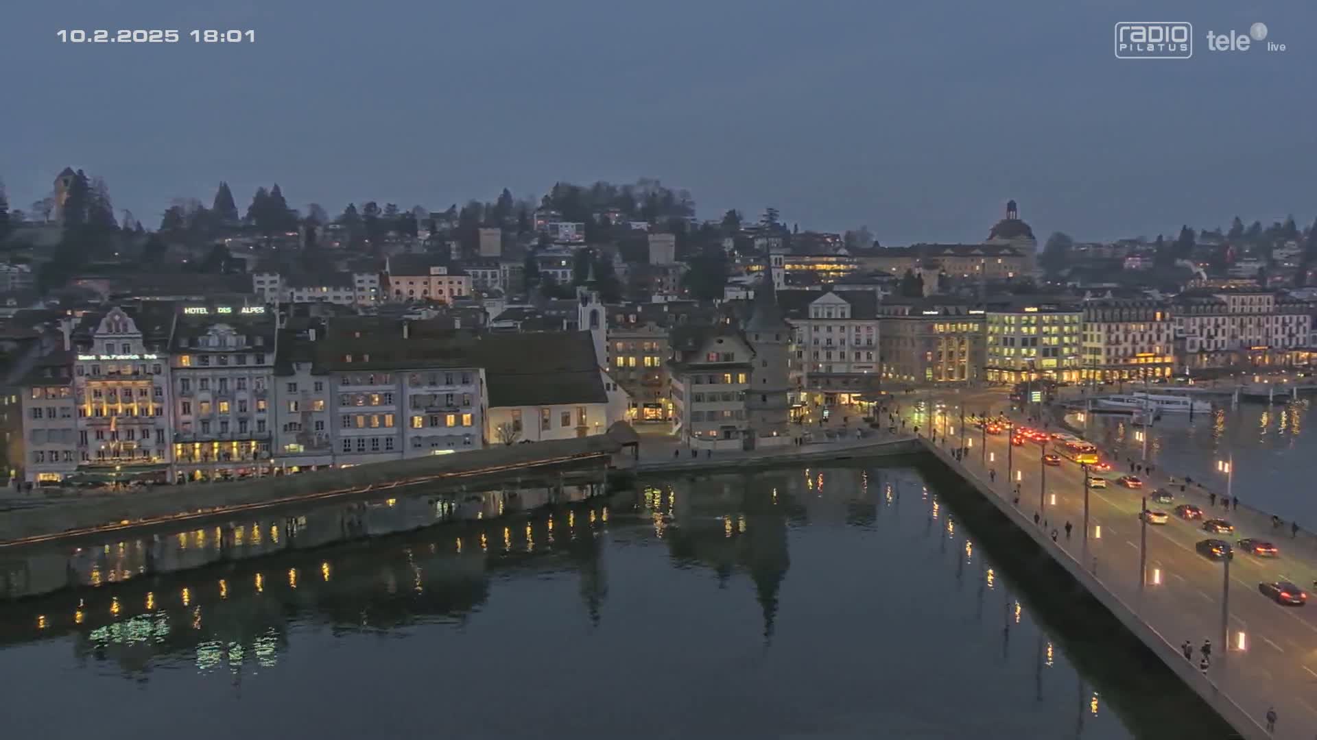A city at twilight is viewed from above, showing a bridge with cars crossing over a dark body of water reflecting city lights.