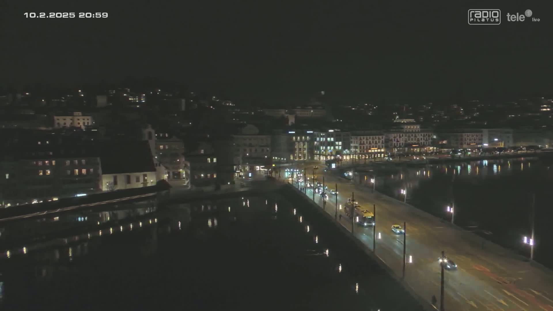 A nighttime, aerial view shows a city's waterfront with a bridge illuminated by streetlights, vehicles traveling on the bridge, and calm water reflecting the city lights.