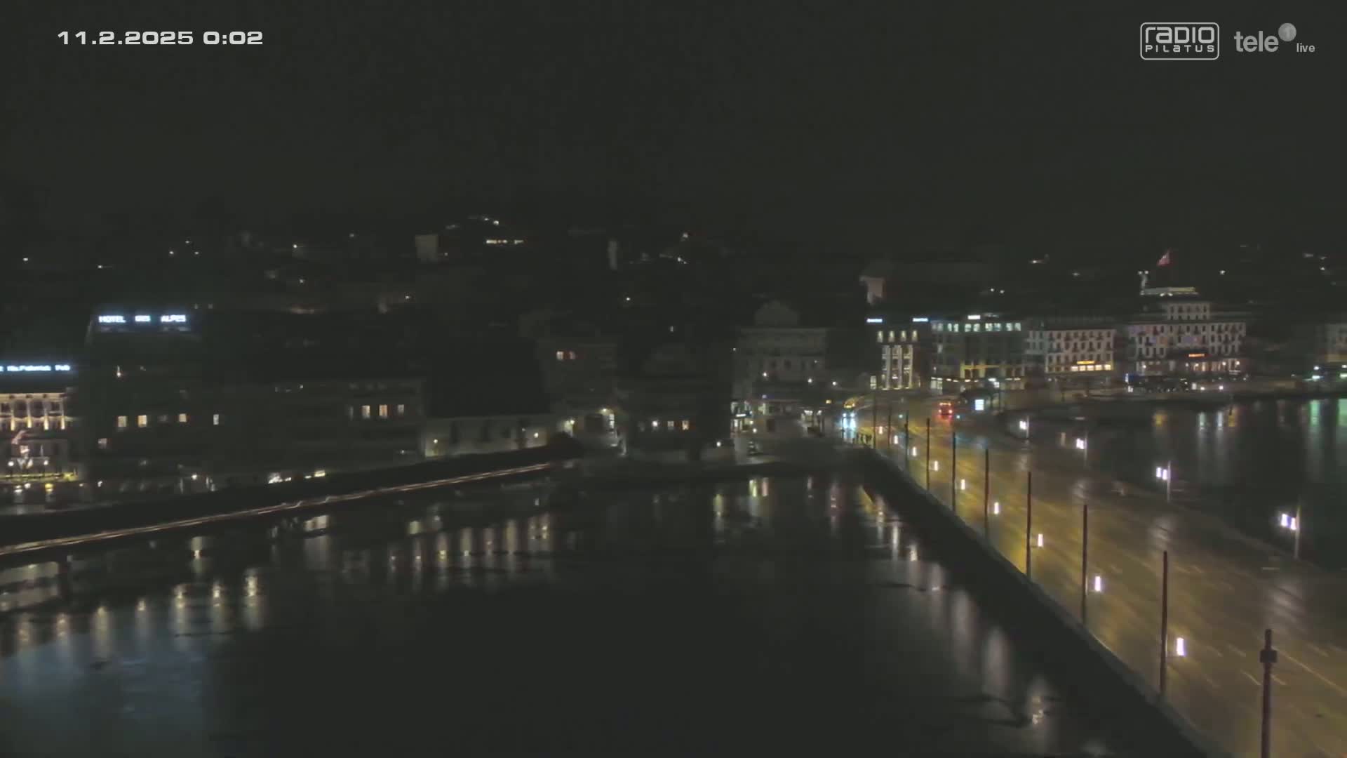 A nighttime, high-angle view shows a city's waterfront with illuminated buildings and a bridge spanning a dark body of water.