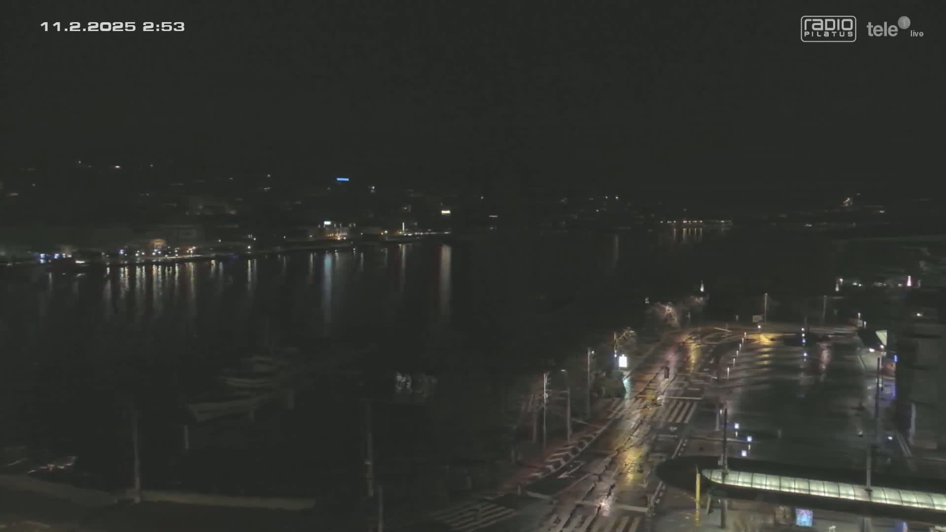 A nighttime view of a city waterfront with a calm body of water reflecting city lights, and a wet, dimly lit street visible in the foreground.