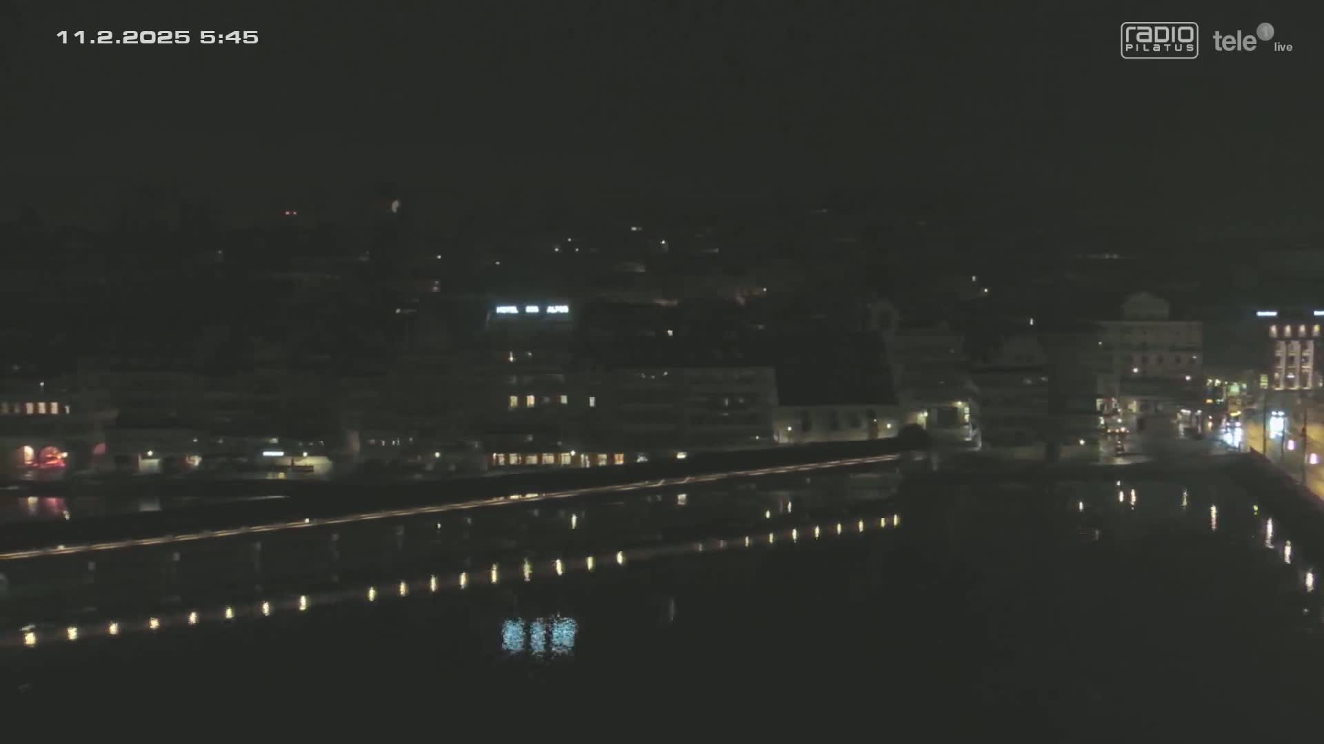 A nighttime, aerial view of a city beside a body of water, showing buildings illuminated with lights reflected on the calm water's surface.