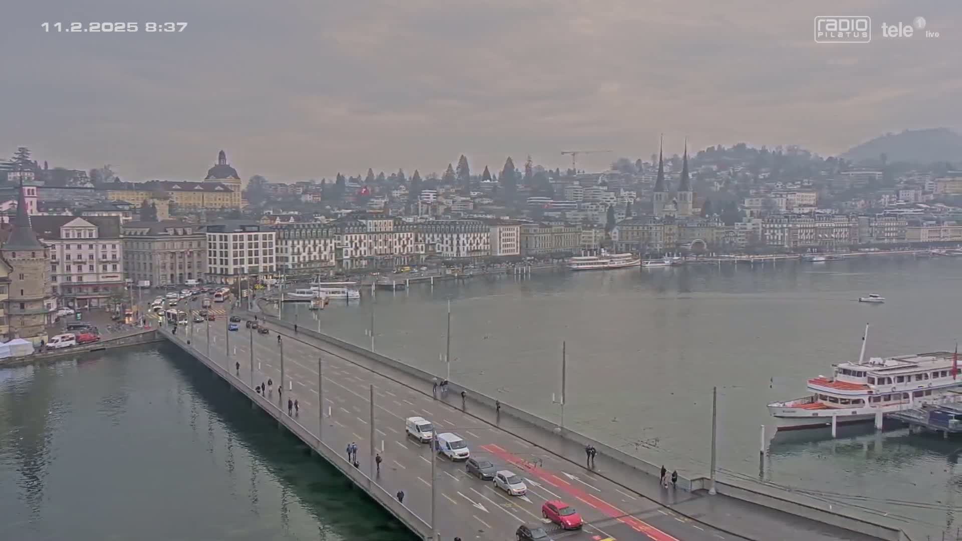 A bridge over a calm body of water leads to a city with overcast skies, where buildings line the waterfront and boats are docked.