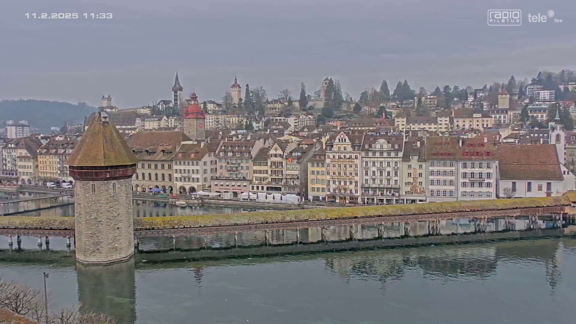 A waterside town with many buildings, a stone tower on a bridge, and overcast skies is reflected in calm water.