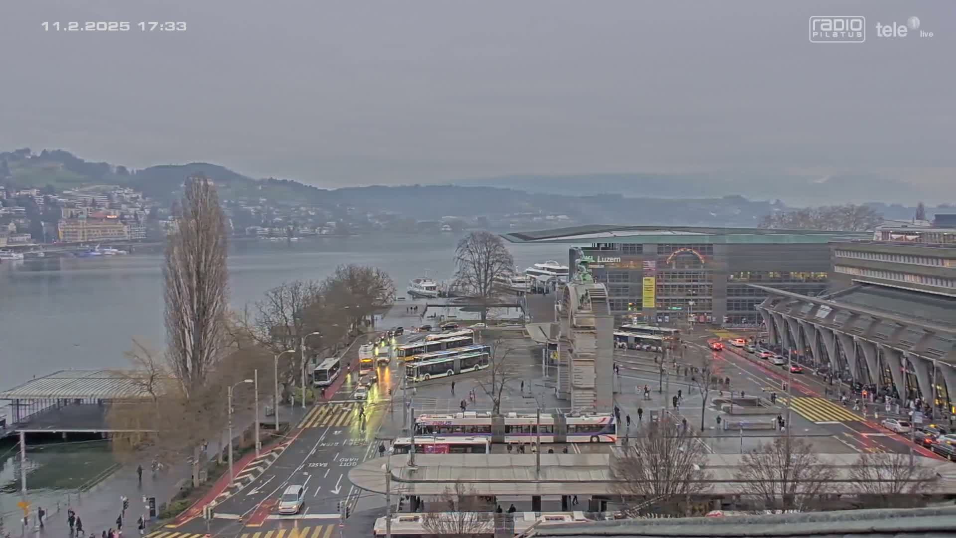 A city waterfront scene on an overcast day shows buses and cars at a transportation hub near a lake with hills in the background.