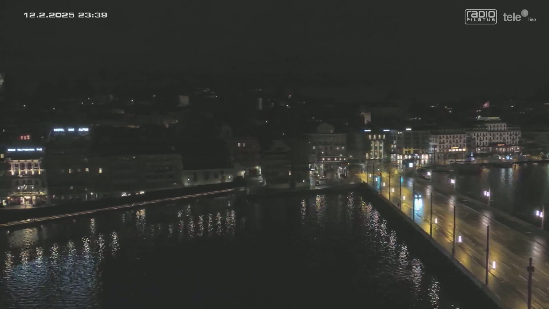 A nighttime aerial view of a city across a body of water, showing a bridge with lights, and buildings illuminated along the water's edge.