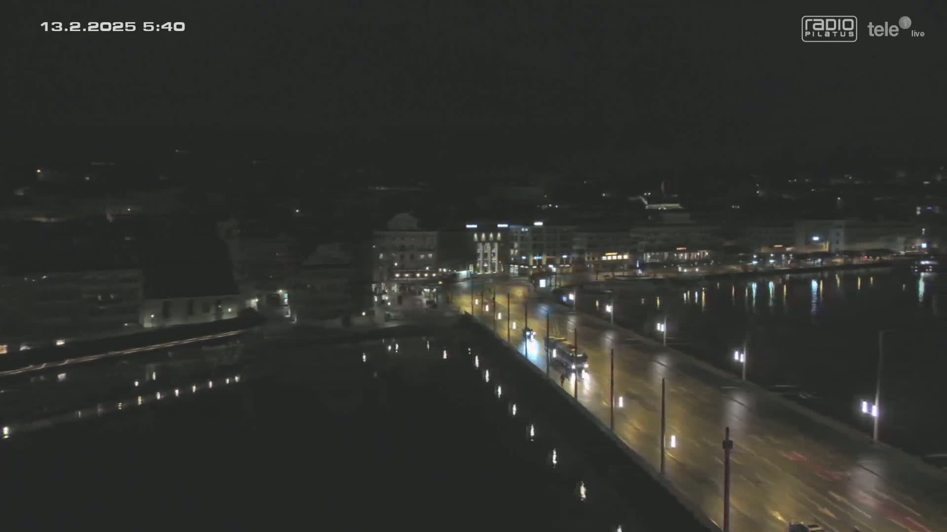 A wet nighttime cityscape is viewed from above, showing a bridge with sparse traffic over dark water reflecting city lights.