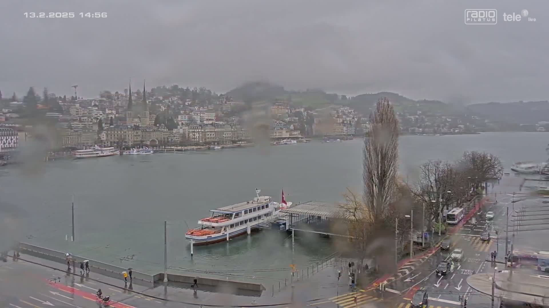 A rainy view of a lake with a docked boat and a city in the background, partially flooded streets and roadways in the foreground.