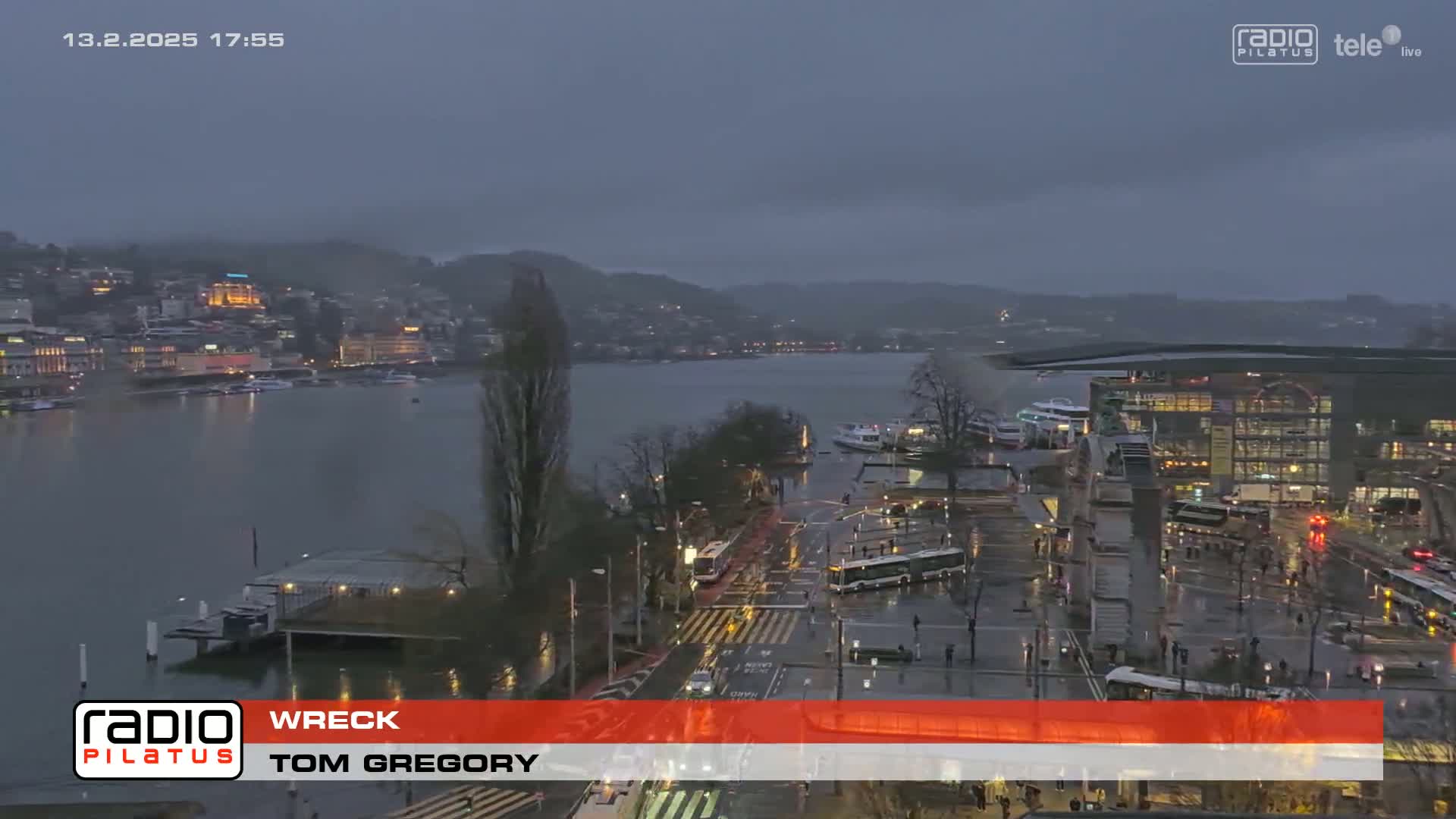 A rain-soaked city waterfront at dusk, showing a body of water with boats and buildings along the shoreline, under a dark, overcast sky.