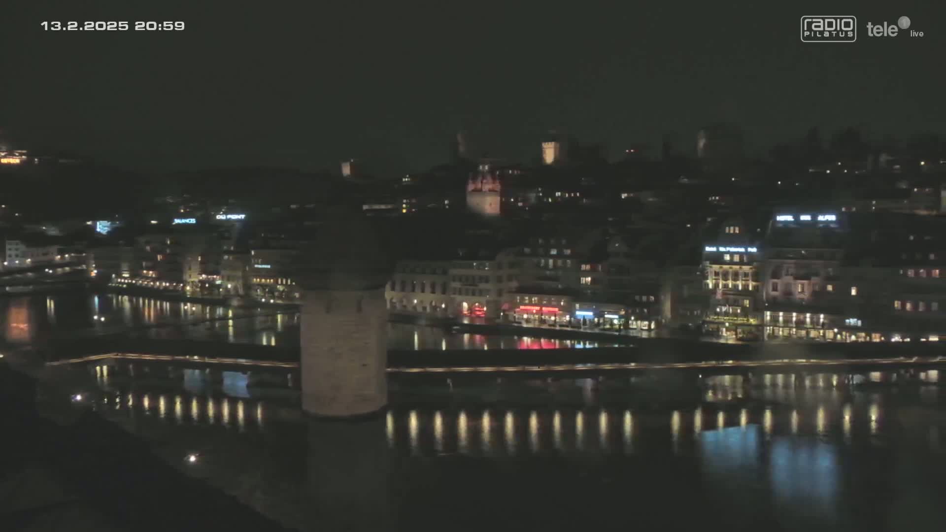 A nighttime view of a city across a calm body of water, showing illuminated buildings and a bridge with a tower.