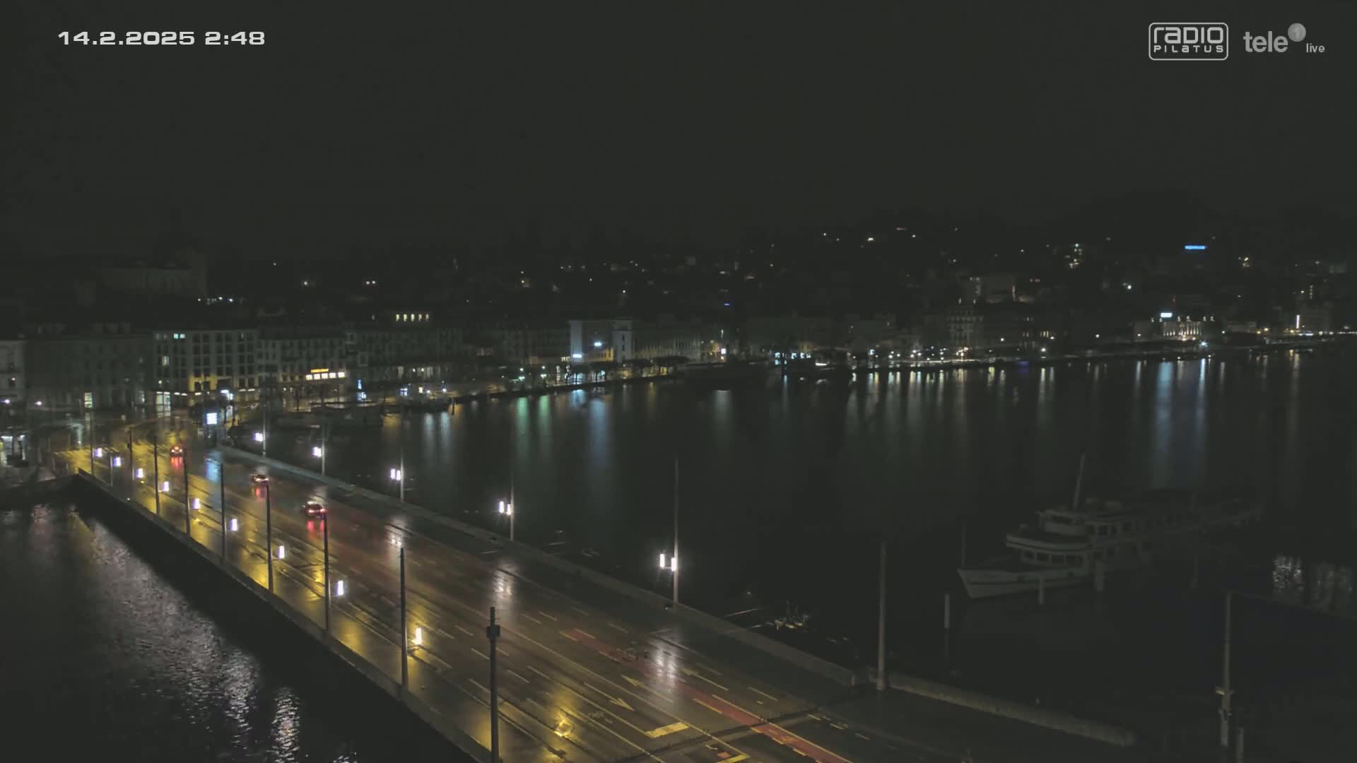 A nighttime view of a wet city bridge with sparse traffic over dark water, alongside a city skyline and a docked boat.