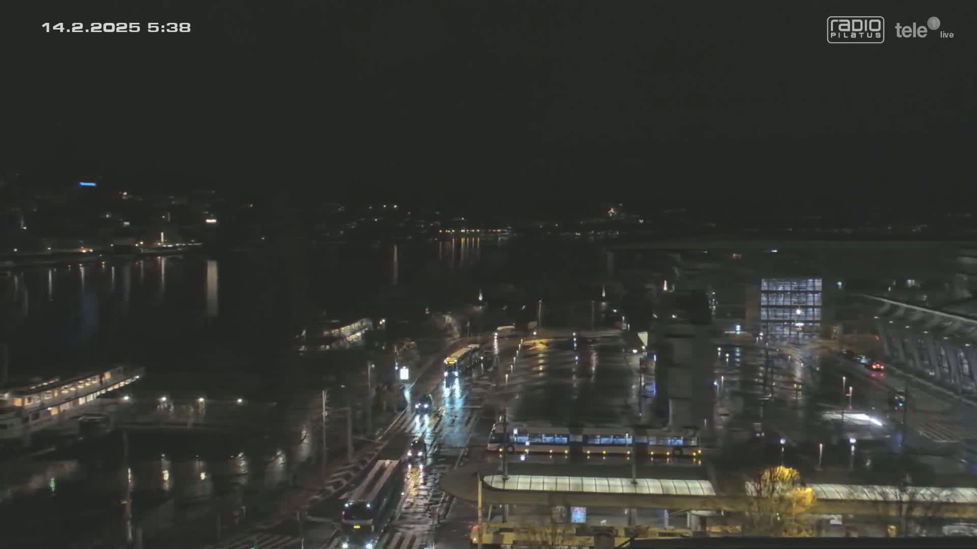 A nighttime, wet city scene shows a body of water reflecting city lights, with trams and vehicles on a roadway near buildings.