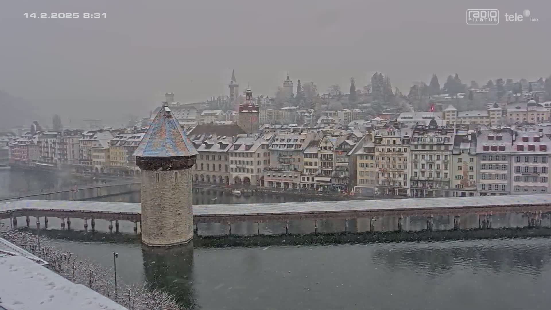 A snow-covered European city with a water channel and a stone tower on a bridge is seen in light snowfall.