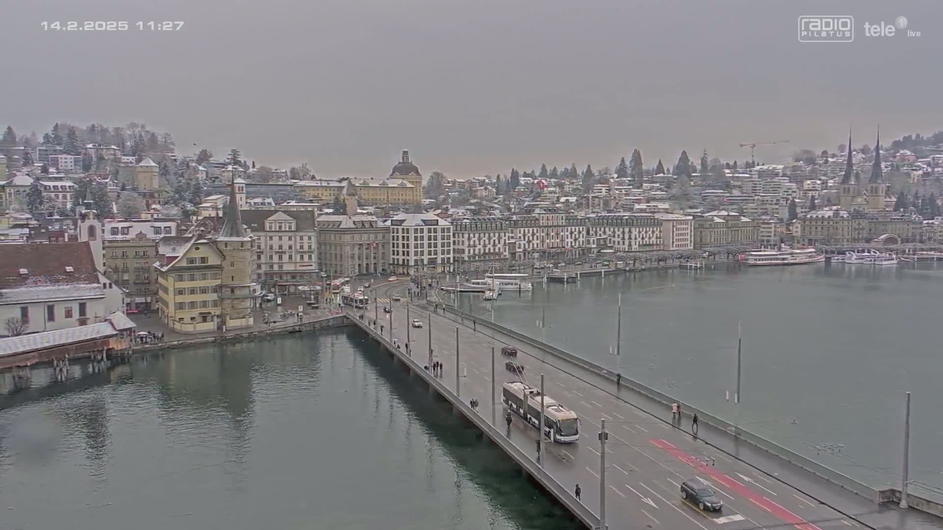A bridge over a calm lake leads to a snow-dusted cityscape under an overcast sky.
