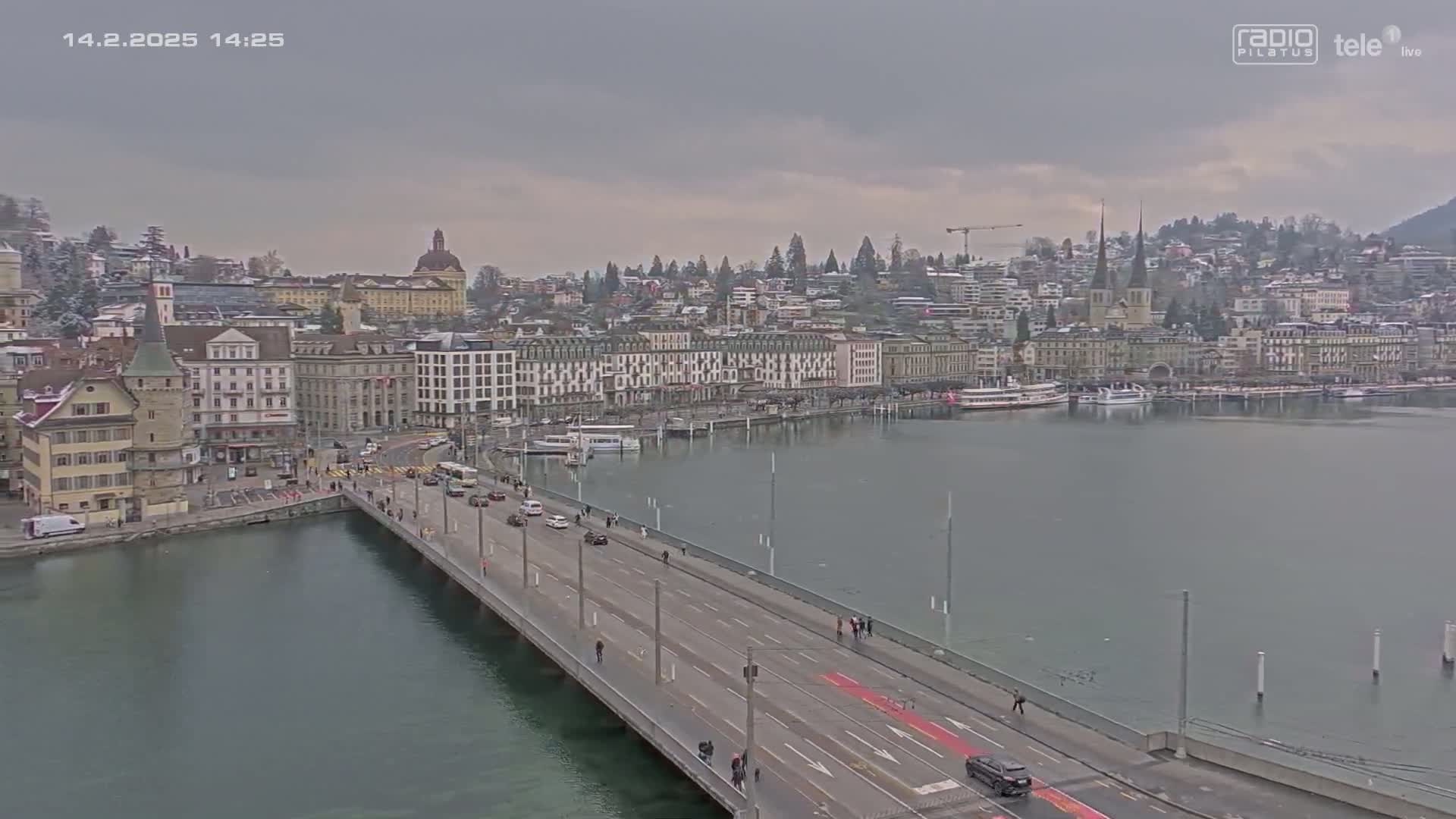 A long bridge with sparse traffic stretches across a calm body of water toward a city of mostly light-colored buildings under an overcast sky.