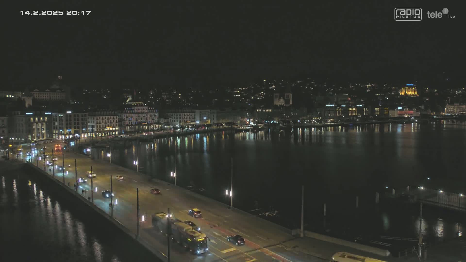 A nighttime view of a city waterfront, showing a bridge with cars driving across it, and buildings lit up along the shore reflecting in the calm water.