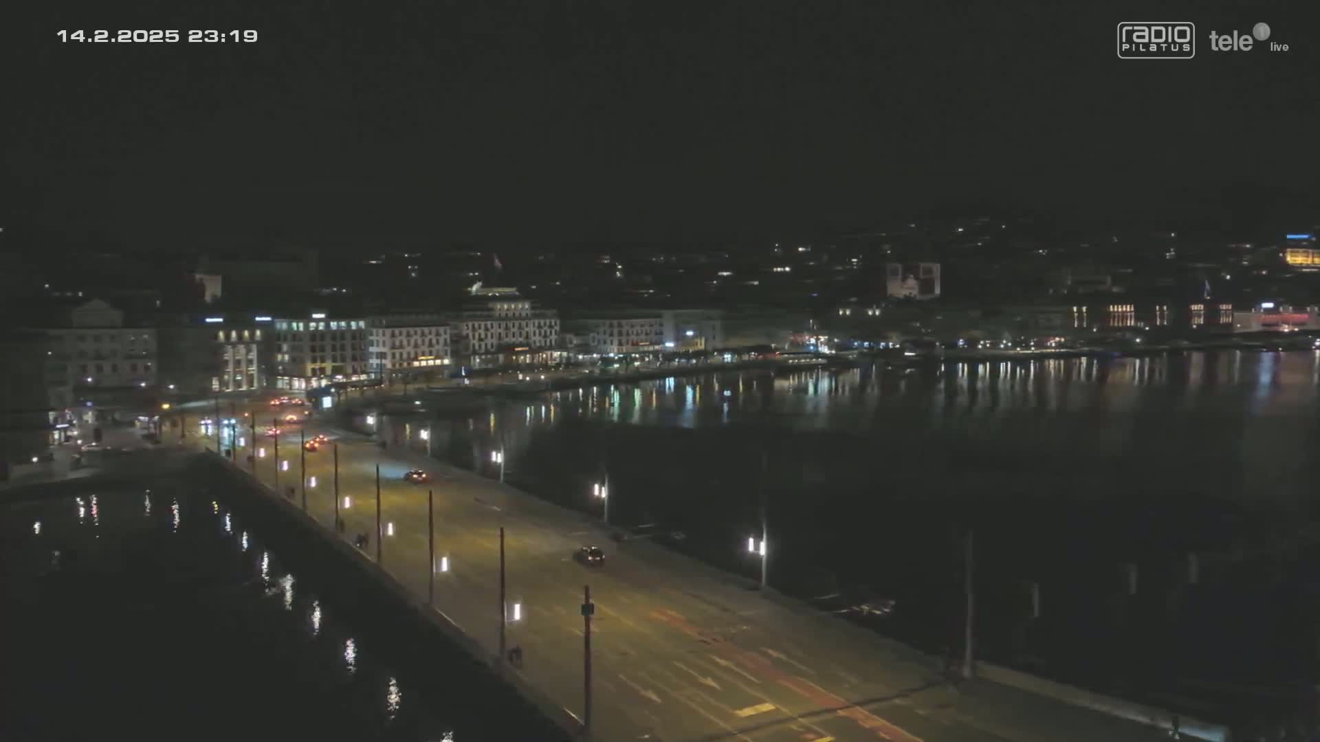 A nighttime, high-angle view shows a city waterfront with a bridge and calm water reflecting the city lights.