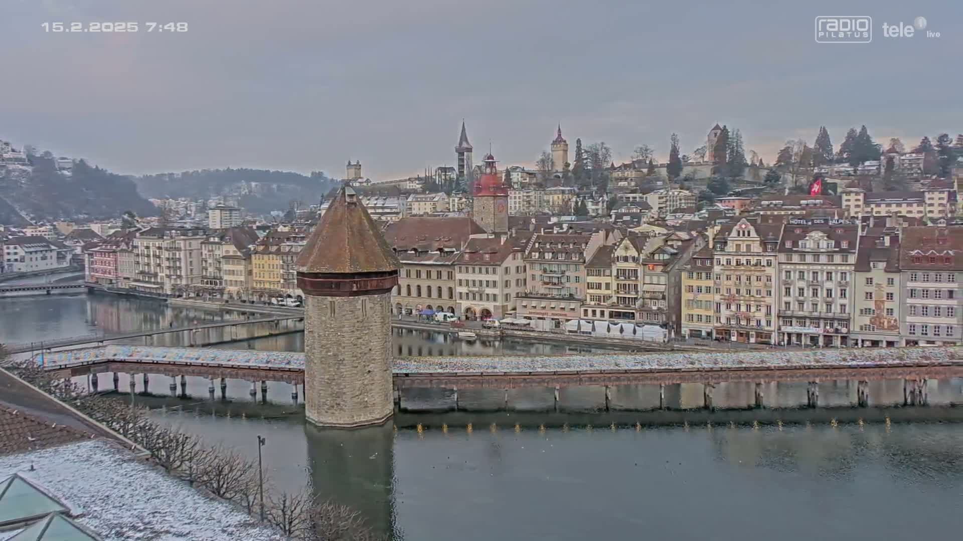 A stone tower stands at the end of a bridge over a calm river, with a town of multi-story buildings and snow-dusted hills in the background under an overcast sky.