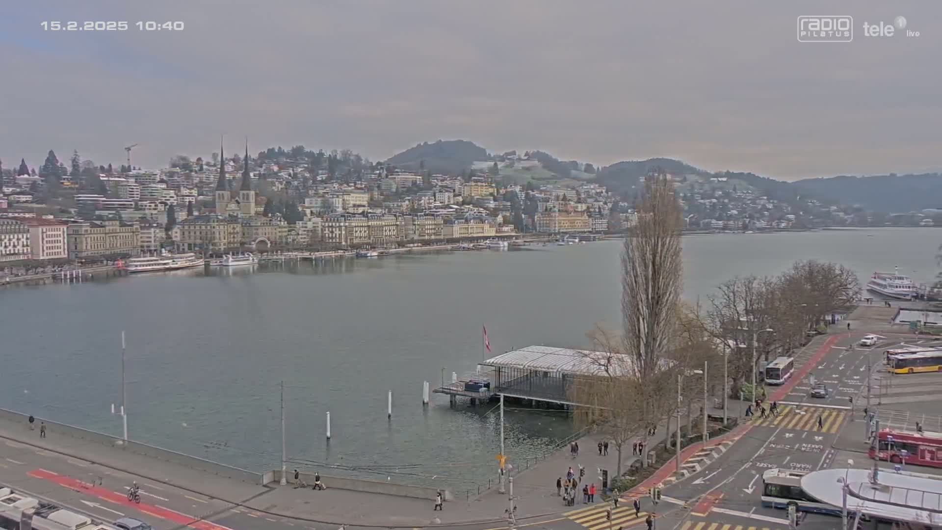 A city waterfront scene on an overcast day shows a body of water with boats and a city skyline of buildings along the shore, with people walking along the waterfront path and buses parked in a nearby lot.