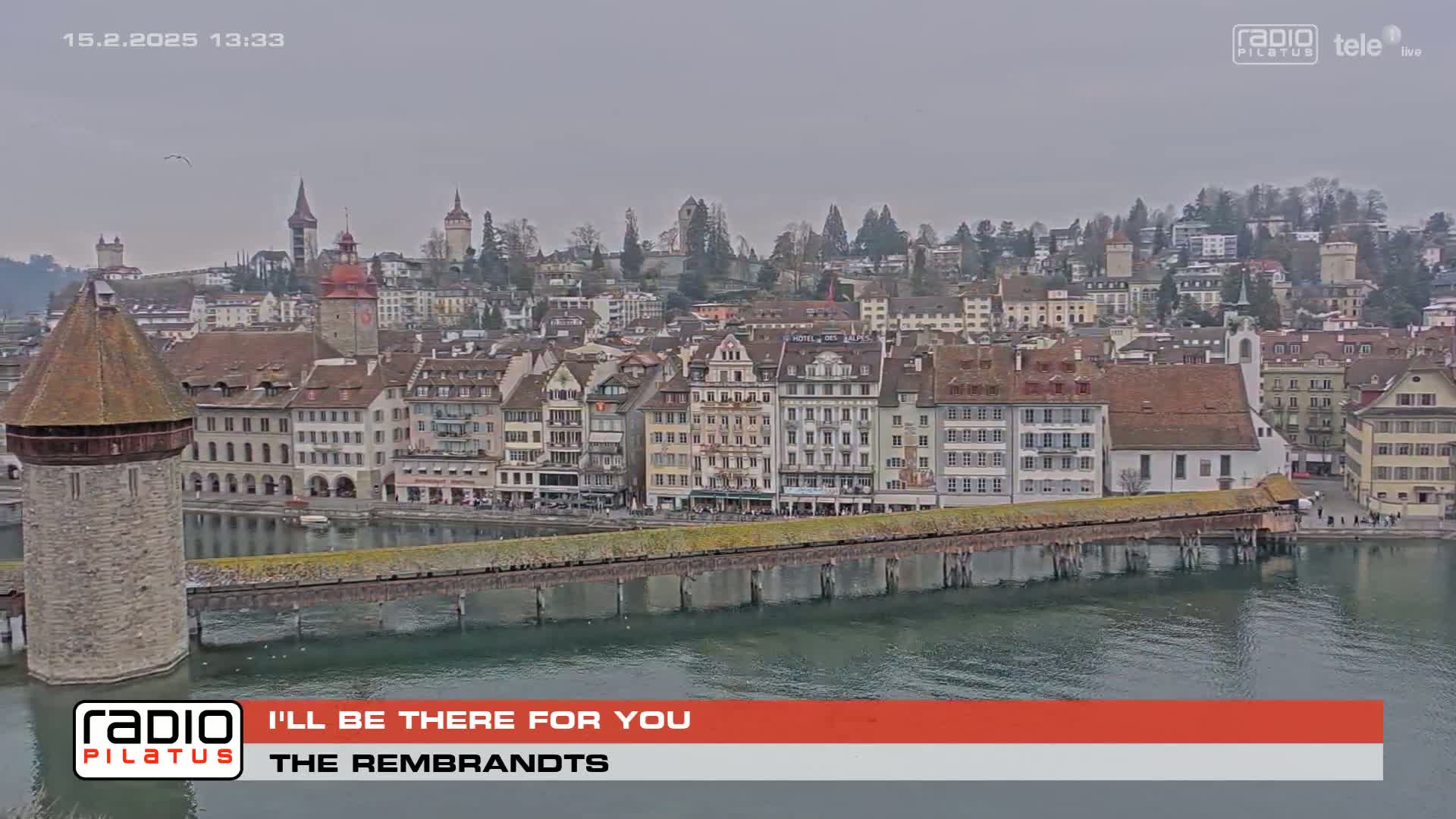 A covered wooden bridge spans a body of water in front of a town of medieval architecture under an overcast sky.
